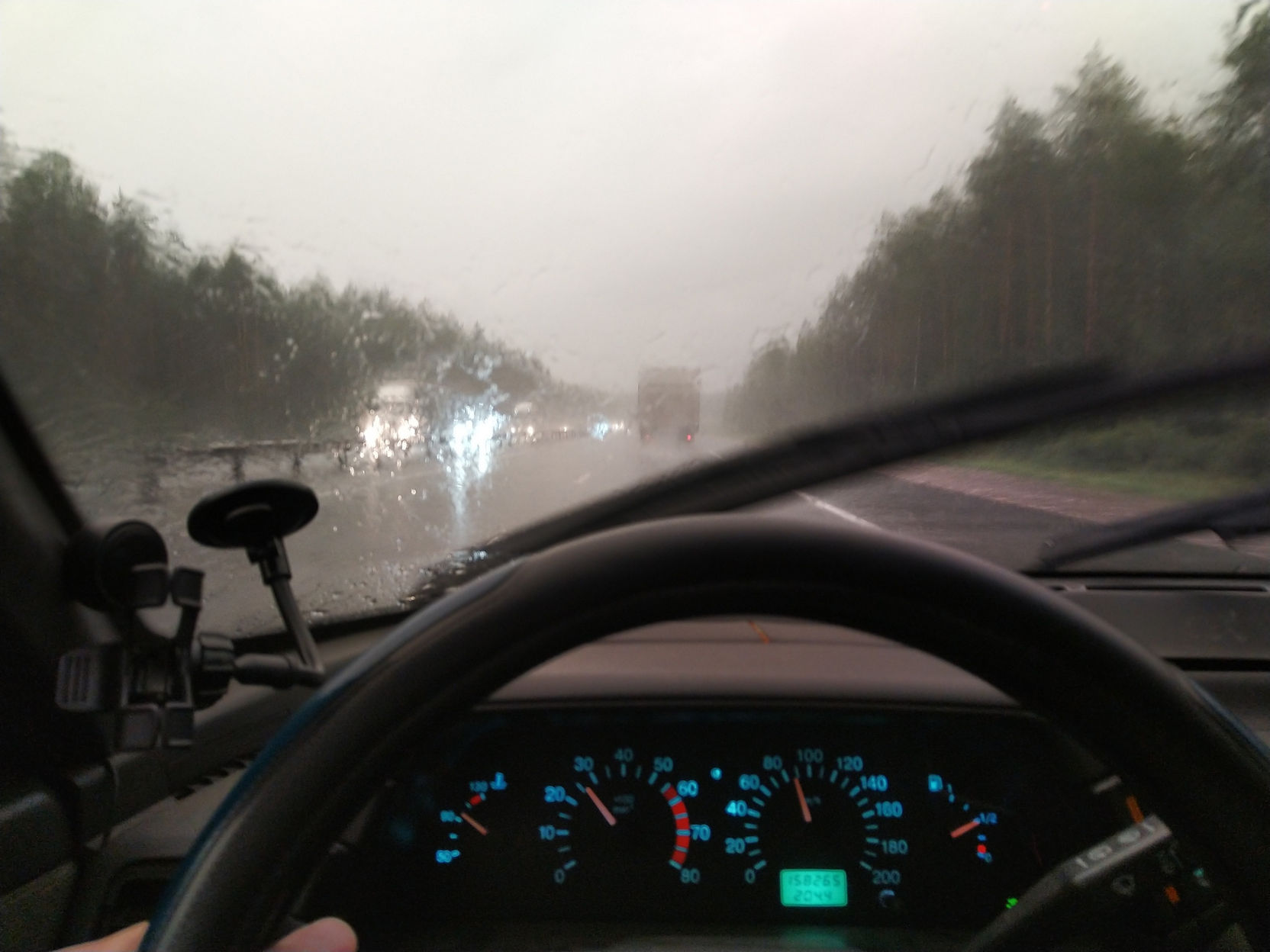 Вид из салона автомобиля через лобовое стекло во время сильного ливня: стекло покрыто крупными каплями дождя, работают дворники; мокрая трасса с деревьями по обочинам, впереди встречные фары и красные огни грузовика; небо светло-серое; руль и приборная панель с синими циферблатами спидометра и тахометра на переднем плане; слева на стекле — держатель навигатора