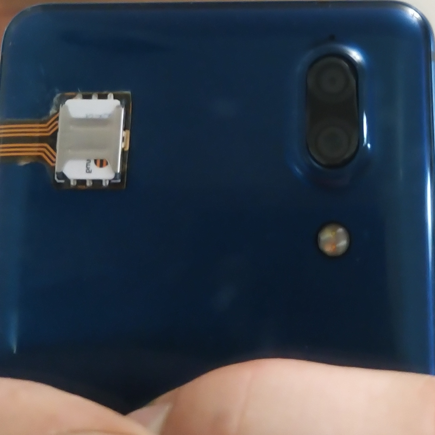 Задняя панель синего смартфона: в левой верхней части корпуса приклеен SIM-адаптер — небольшое белое устройство с оранжевым гибким шлейфом, уходящим под корпус телефона; в правом верхнем углу — двойная камера; телефон держат в руке снизу