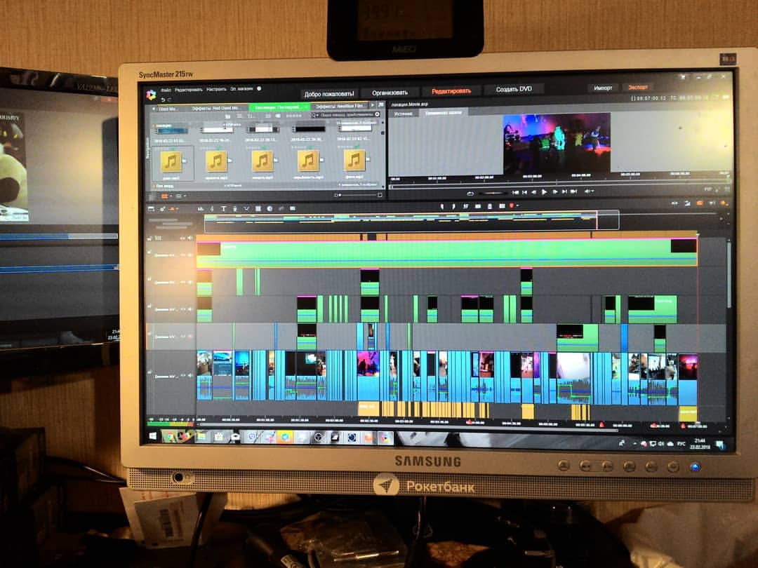Монитор Samsung SyncMaster со стикером Рокетбанк: на экране — программа Pinnacle Studio с длинным проектом; таймлайн заполнен разноцветными клипами на множестве видео- и аудиодорожек; в правом верхнем углу — превью кадра с концертом; слева виден второй монитор NEC