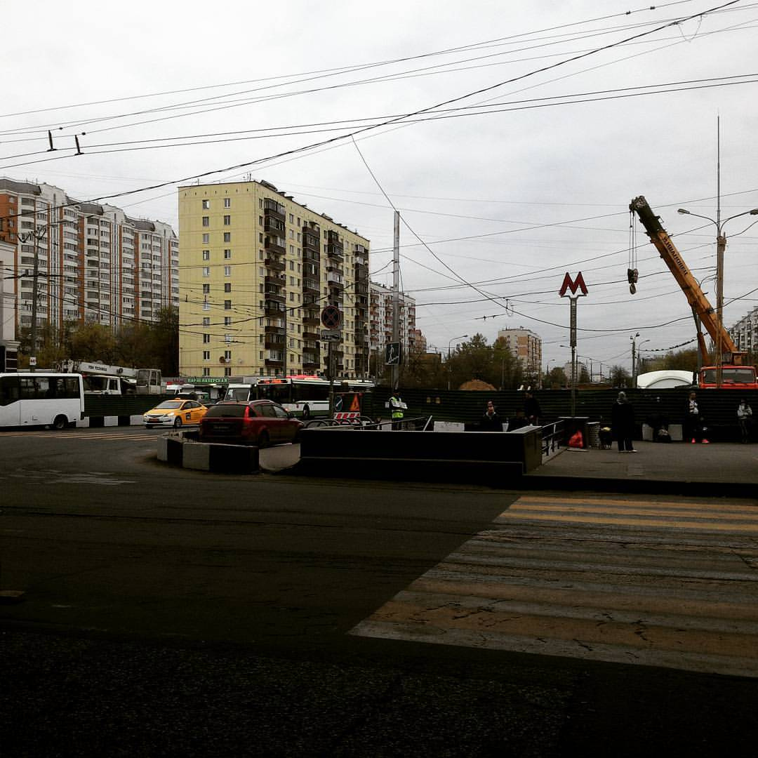 Московская улица у станции метро (красная вывеска «М» видна справа): большая строительная площадка с зелёными металлическими ограждениями, жёлтый строительный кран, автобус, такси, пешеходы; над дорогой — троллейбусные провода; на фоне — многоэтажные панельные дома, пасмурное небо