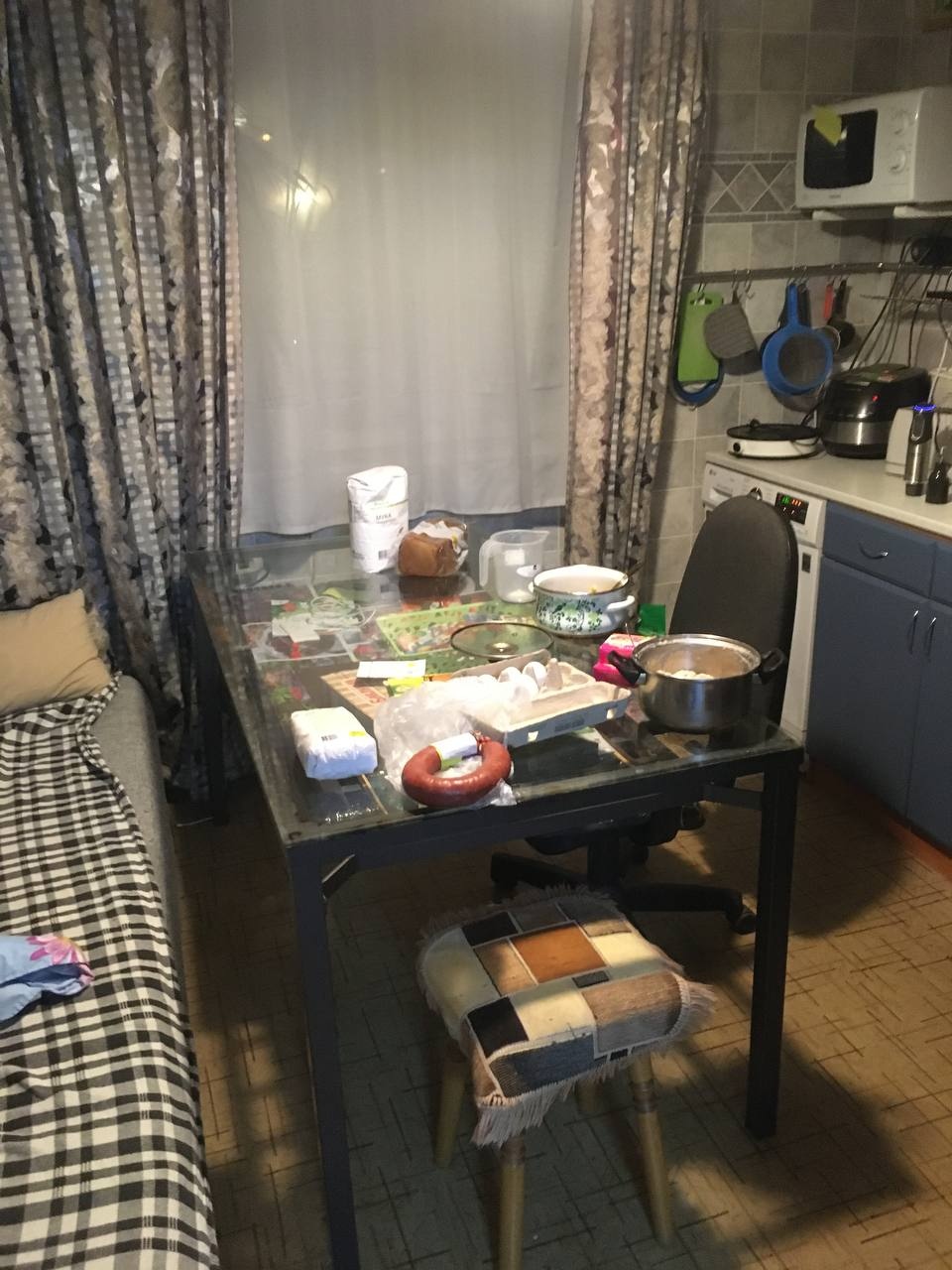 📷 Небольшая кухня: стол со стеклянной столешницей, на нём посуда, кастрюля, еда в беспорядке, холодильник, занавески с рисунком, стул с тканевой обивкой.