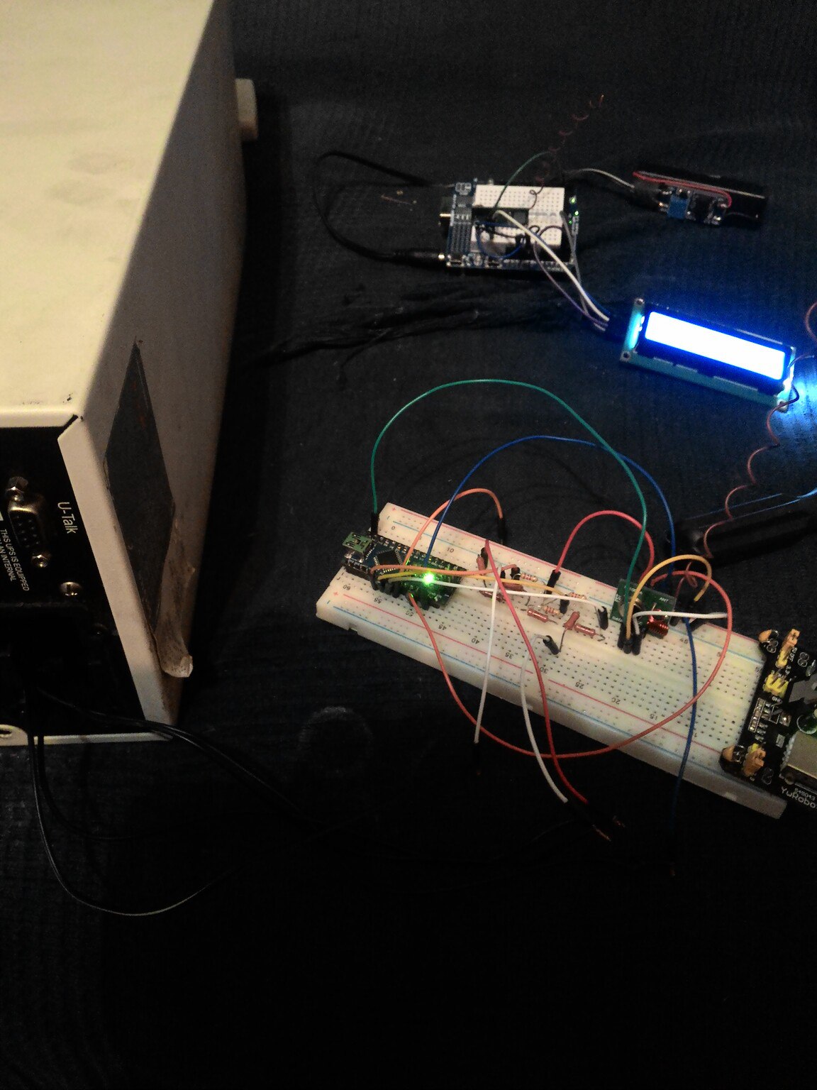 Фото сверху: на тёмной поверхности макетная плата с Arduino Nano, блоком питания и компонентами; в верхнем правом углу синий LCD-дисплей 16×2 с подсветкой; рядом белый картон/коробка