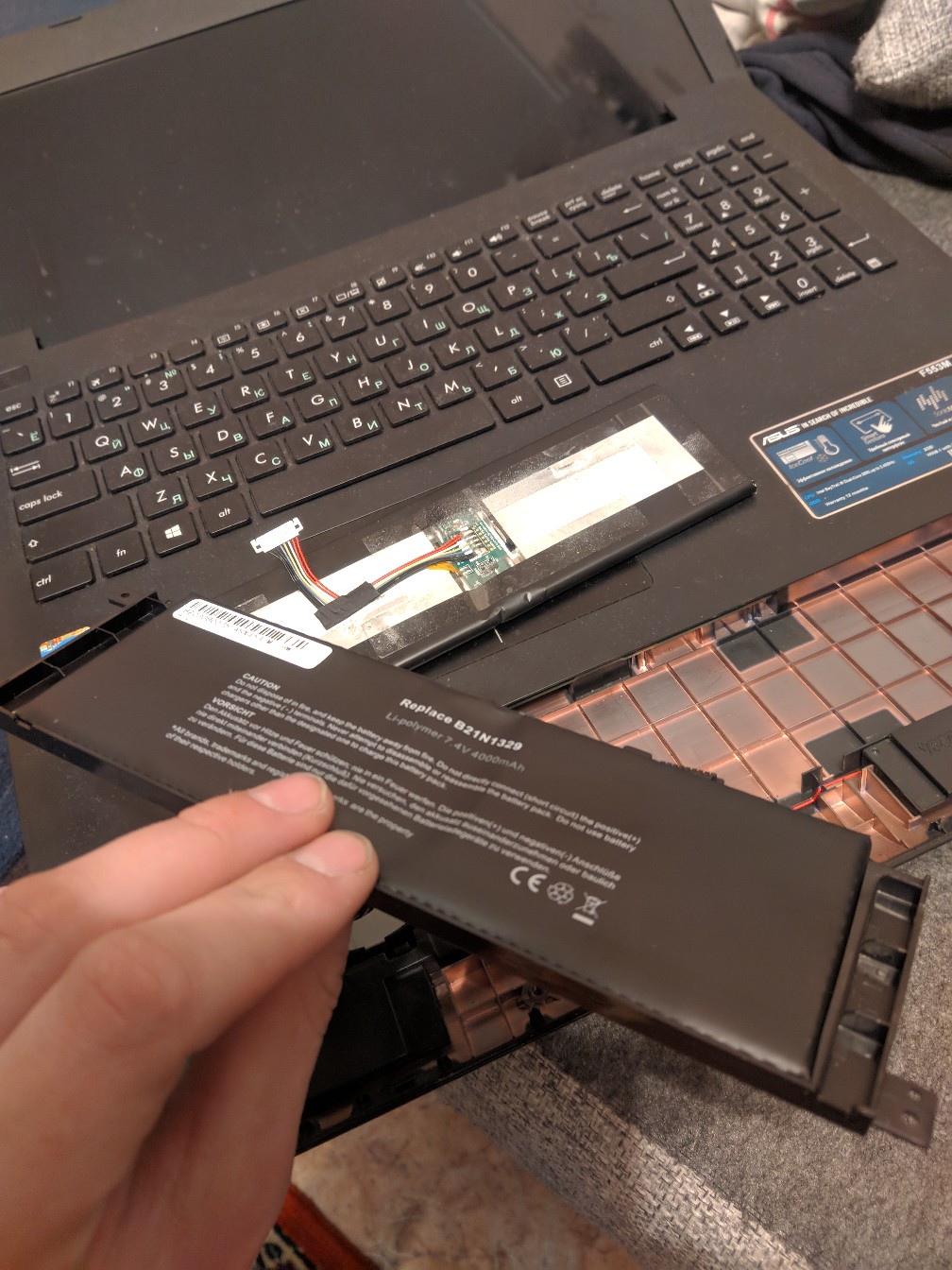 Ноутбук ASUS (чёрный корпус) открыт; рука извлекает тонкий чёрный аккумулятор с белой наклейкой; видна клавиатура и внутренний разъём батареи