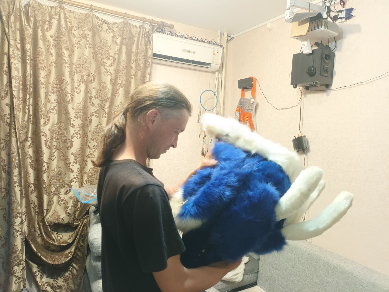 📷 Человек с длинными светлыми волосами держит синюю фурри-голову (волк/пёс) в комнате с кондиционером на стене.
