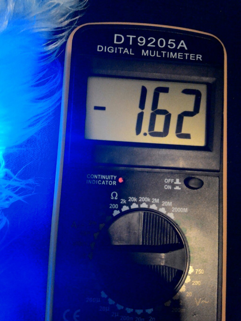 Крупный план мультиметра DT9205A Digital Multimeter: на дисплее «−1.62»; переключатель режимов повёрнут в сторону сопротивления (диапазон около 2k Ω); красный индикатор Continuity; синяя подсветка фона, рядом слева видно что-то пушистое белое