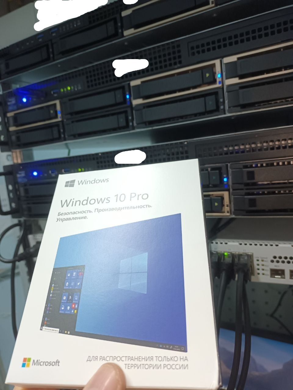 📷 Рука держит коробку Microsoft Windows 10 Pro («Безопасность. Производительность. Управление. / ДЛЯ РАСПРОСТРАНЕНИЯ ТОЛЬКО НА ТЕРРИТОРИИ РОССИИИ») на фоне серверной стойки с несколькими серверами и синими индикаторами.