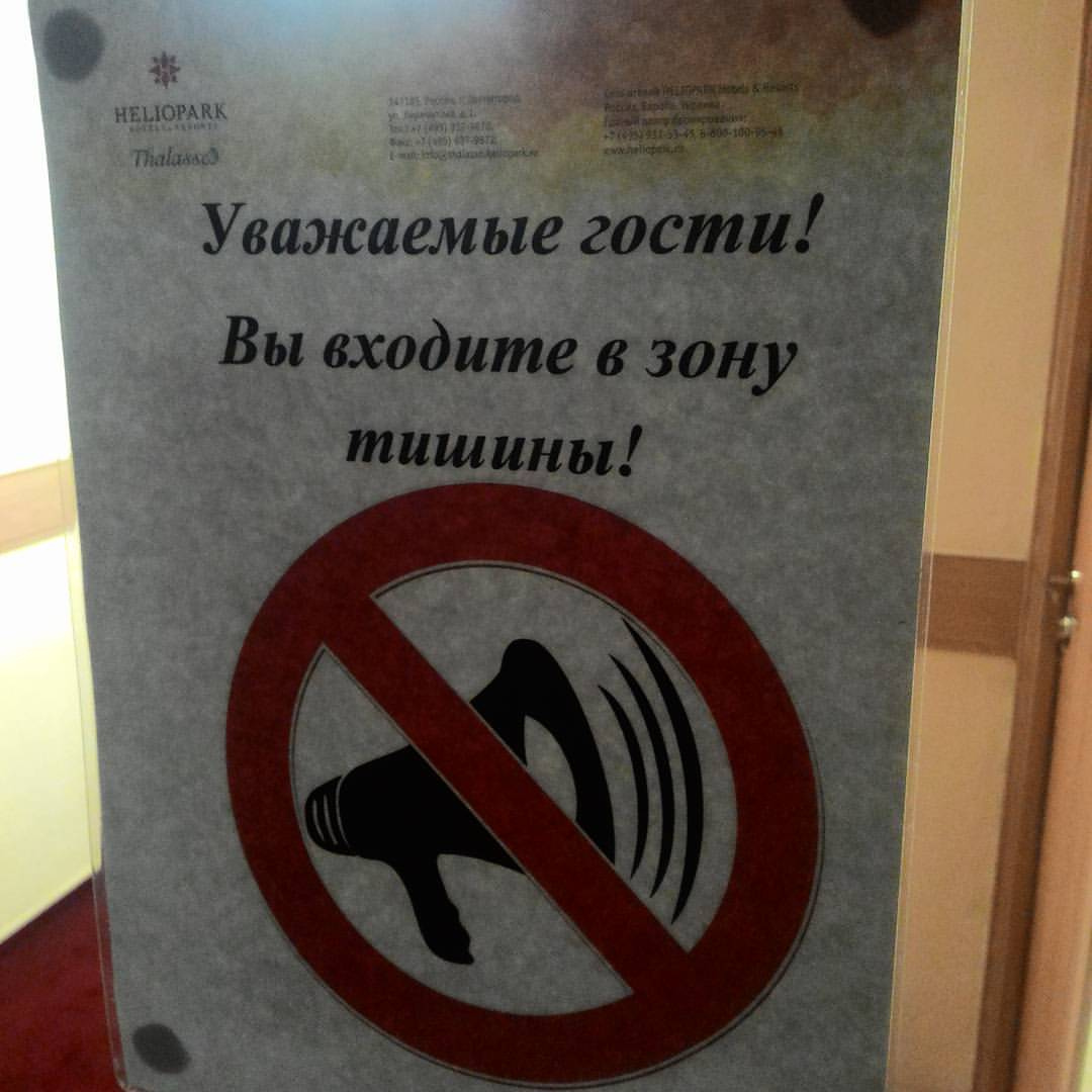 Табличка на двери: в верхнем левом углу — логотип «HELIOPARK» со снежинкой и подпись «ThalaNes3»; в центре крупным шрифтом: «Уважаемые гости! / Вы входите в зону тишины!»; ниже — большой красный знак запрета шума (красный круг с перечёркнутым динамиком)