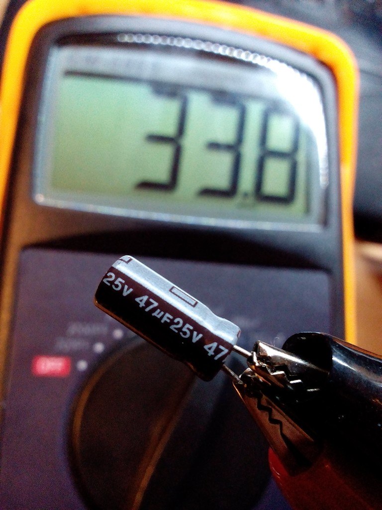 Жёлто-чёрный мультиметр показывает 33.8 мкФ; на щупах зажат электролитический конденсатор 47 мкФ 25В — ёмкость занижена, конденсатор высох