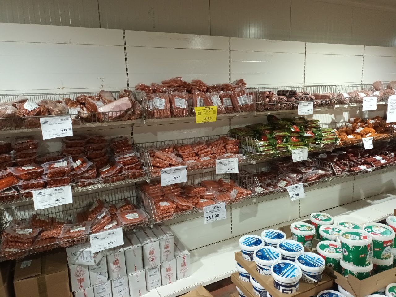 📷 Полки холодильника с мясными продуктами: сосиски, колбасы разных видов, ценники на полках; справа — белые крышки (йогурт/творог).