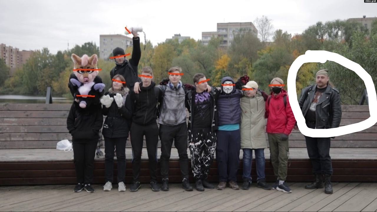 📷 Групповое фото на деревянном мосту/набережной: ~9 человек стоят в ряд, обнявшись; крайний слева в голове фурсьюта (свиноподобный персонаж); лица остальных закрыты оранжевыми прямоугольниками; у одного поднята рука с чем-то оранжевым; справа в белом круге выделен мужчина в чёрной куртке; осенние деревья и многоэтажки на фоне.