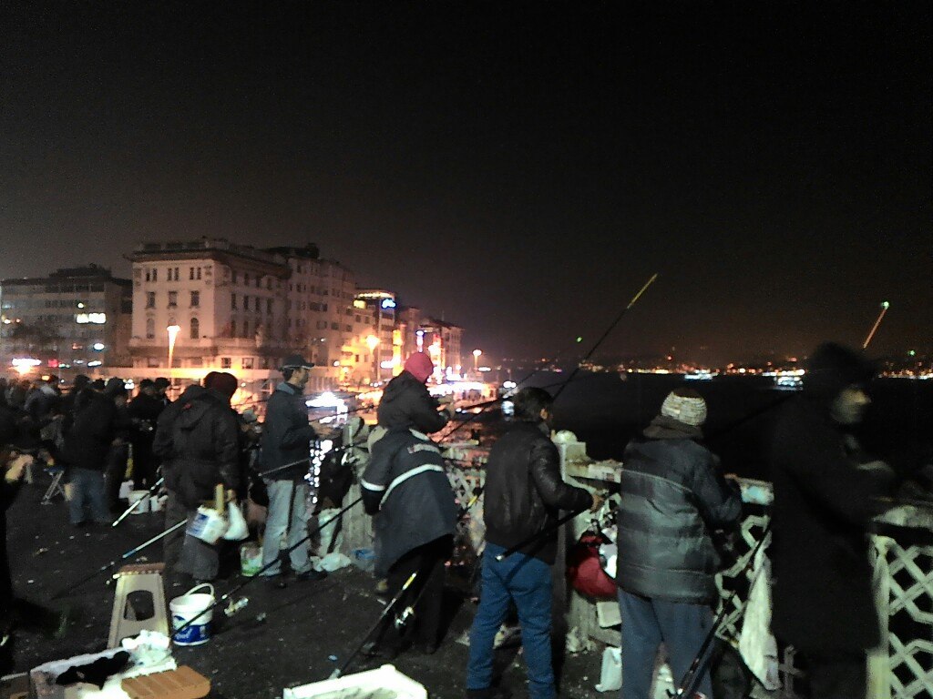 Рыбаки ночью на Галатском мосту: плотный ряд мужчин с удочками, закинутыми в Золотой Рог; вёдра и снасти на перилах; вдали — подсвеченная Новая мечеть (Yeni Cami) с двумя минаретами и набережная Эминёню