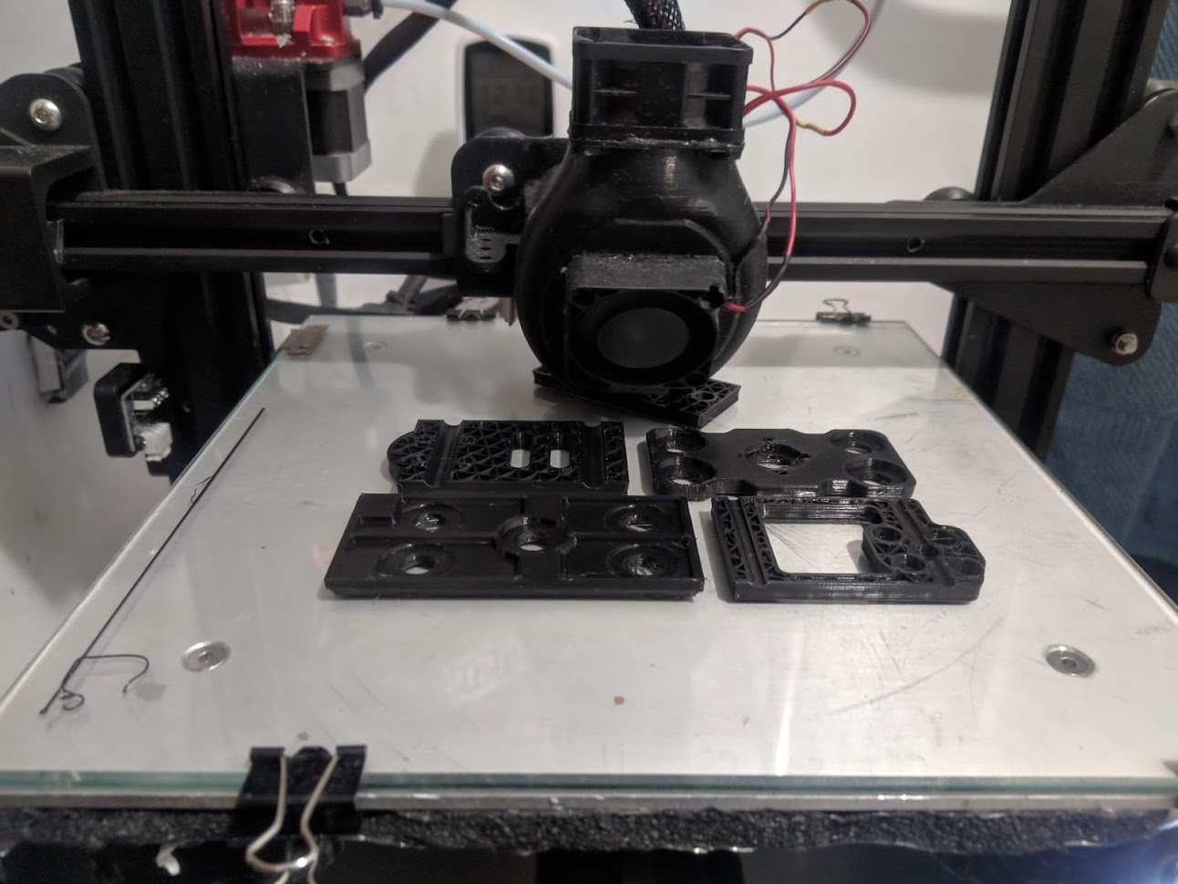 📷 Стол 3D-принтера с двумя напечатанными чёрными пластиковыми деталями (кронштейны/корпуса виброразвязки); чёрная металлическая рама принтера на фоне, белая рабочая платформа.