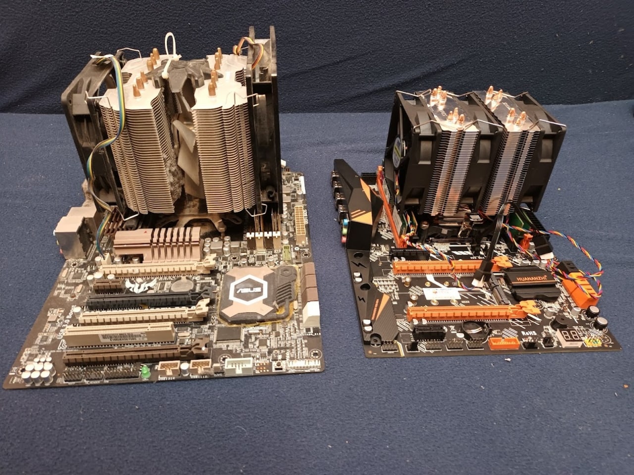 📷 Две материнские платы ATX рядом на поверхности, каждая с башенным кулером с вентилятором; левая — тёмный PCB, правая — с оранжевыми акцентами.