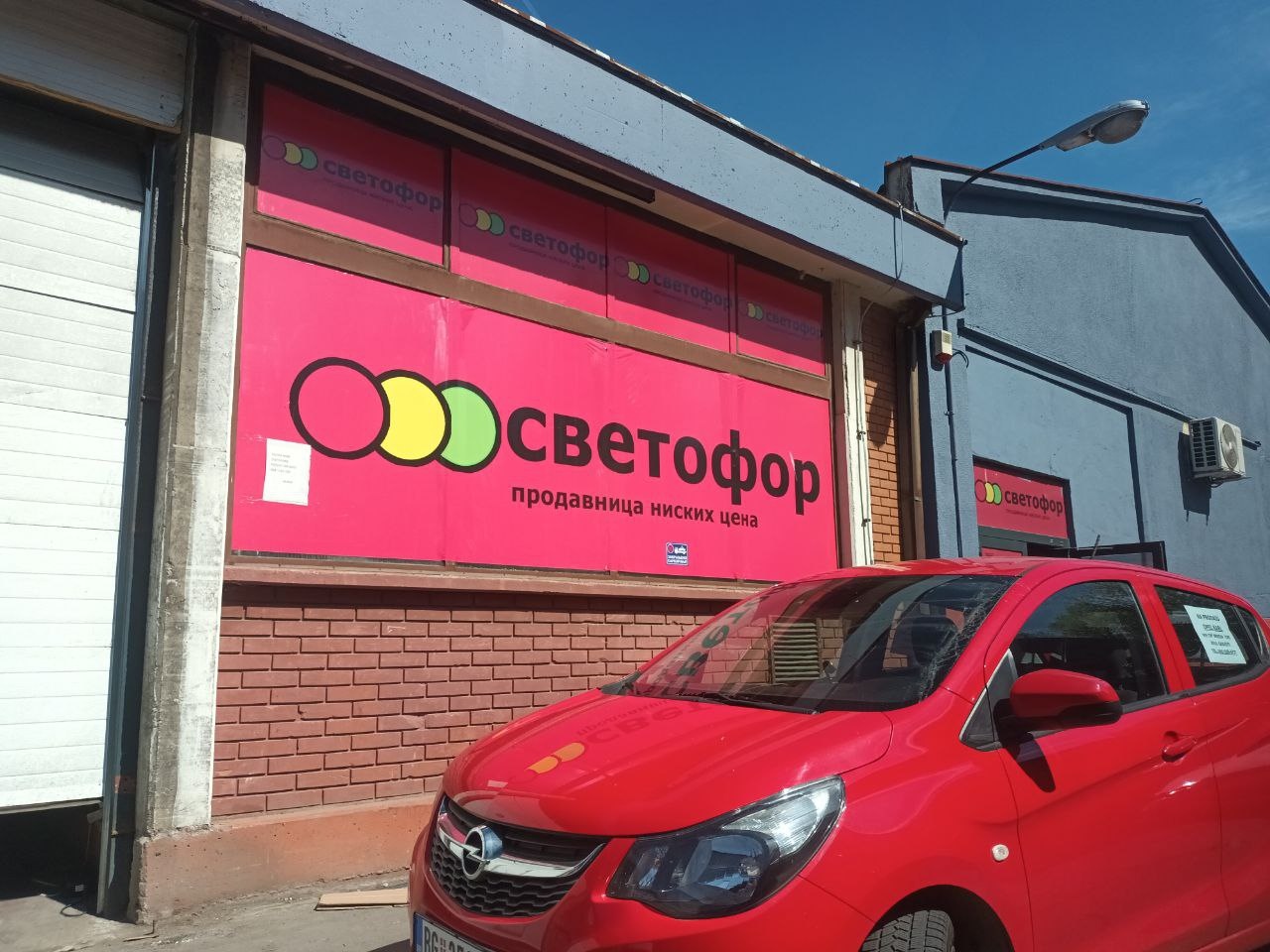 📷 Фасад магазина «Светофор — продавница ниских цена»: красно-оранжевая вывеска с логотипом (три кружка — зелёный, жёлтый, красный); перед входом — красная машина Opel.
