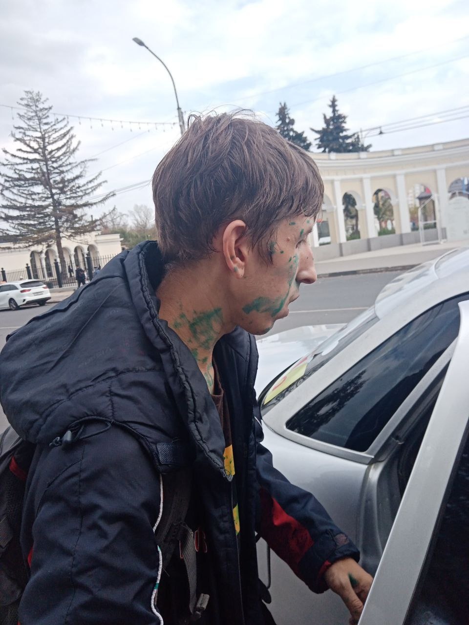 📷 Молодой человек с зелёнкой на лице стоит у серебристого автомобиля на улице; на заднем плане — деревья, нарядная арка, здание.
