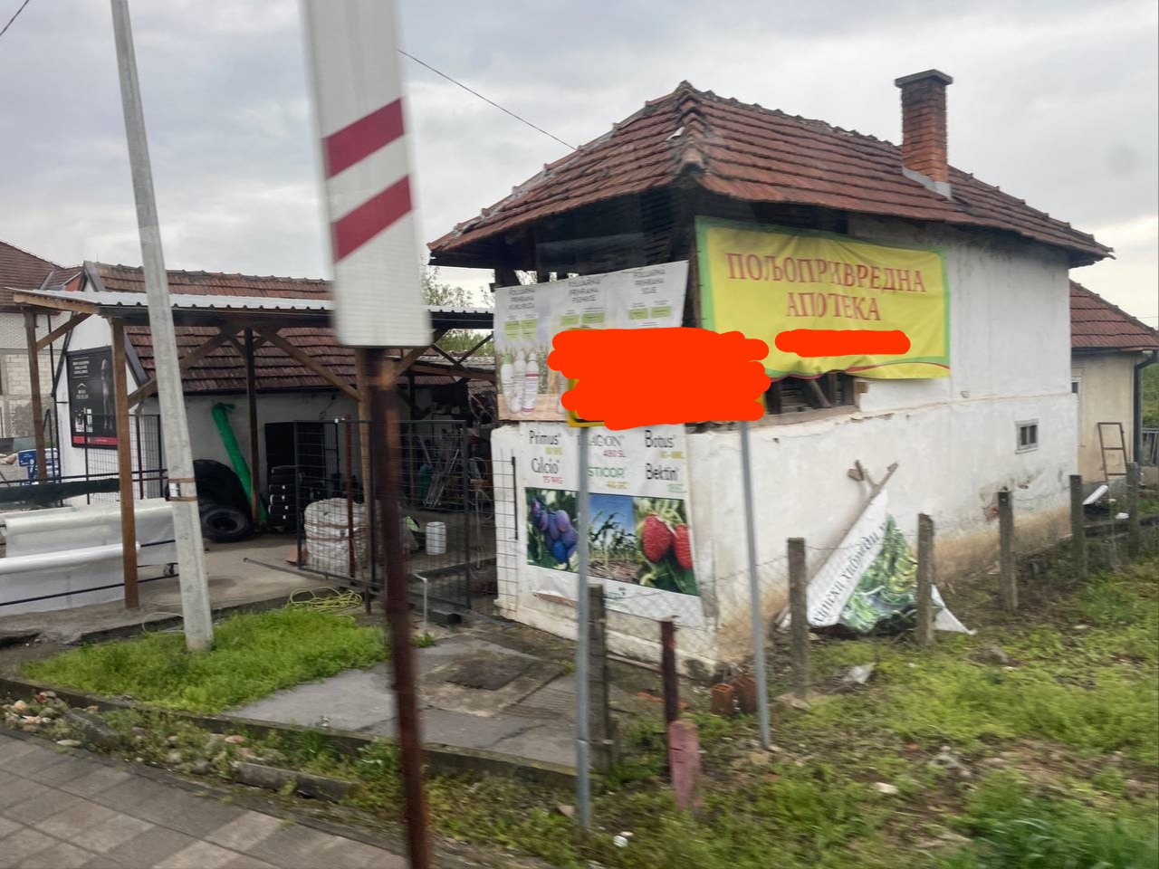 📷 Сербия: небольшое белое здание «ПОЉОПРИВРЕДНА АПОТЕКА» (сельскохозяйственная аптека) с жёлто-зелёной вывеской; деревья, пасмурно.