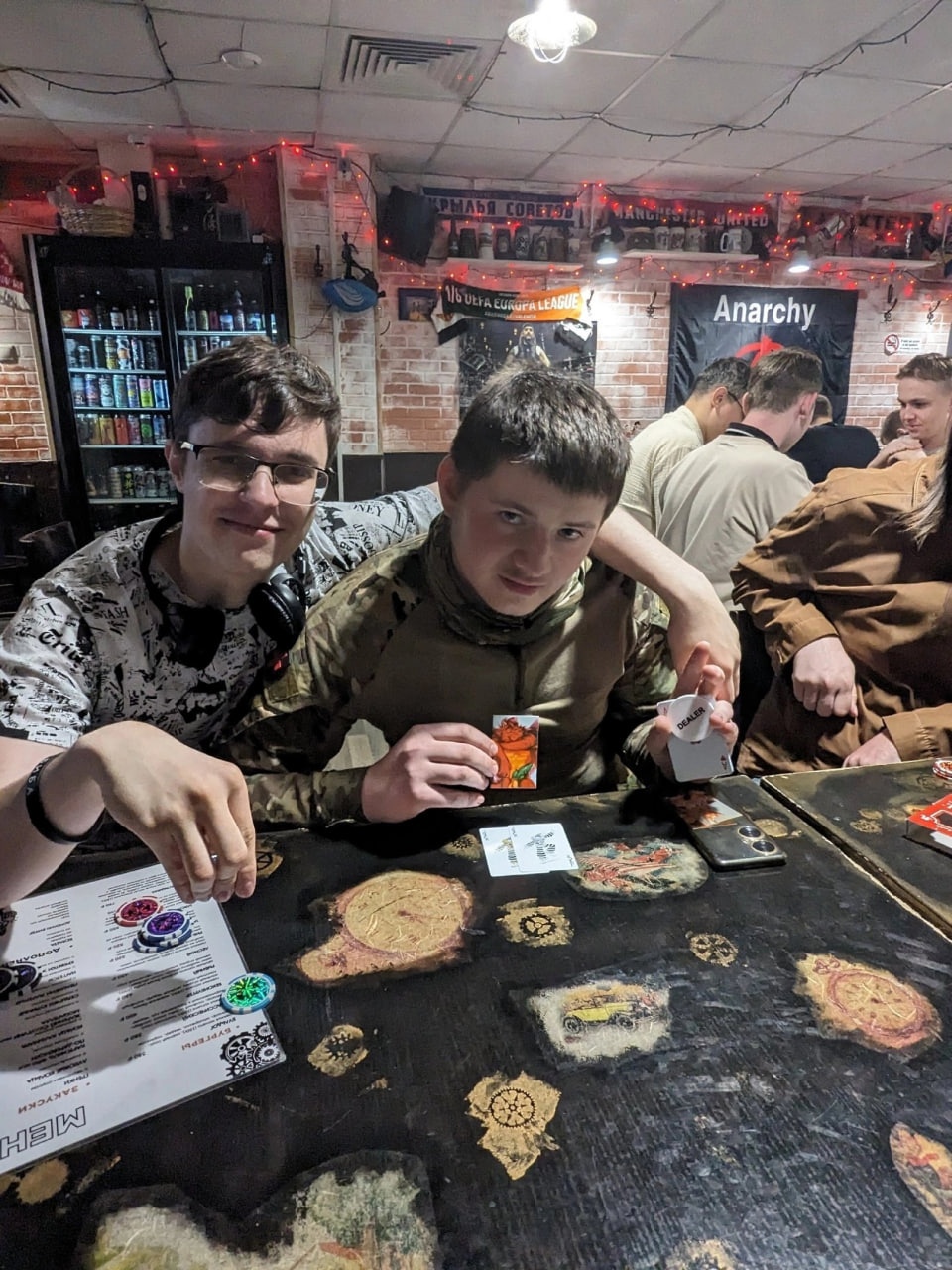 📷 Двое молодых людей за столиком в тёмном баре с надписью «Anarchy», играют в настольную игру.