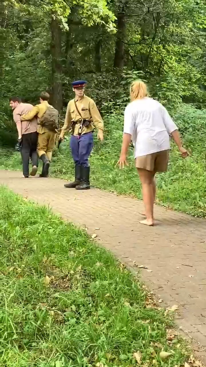 📷 Трое идут по дорожке в летнем парке: мужчина в форме советской/сталинской милиции (синий китель, фуражка, портупея, сапоги), следом двое в обычной одежде; зелёные деревья.