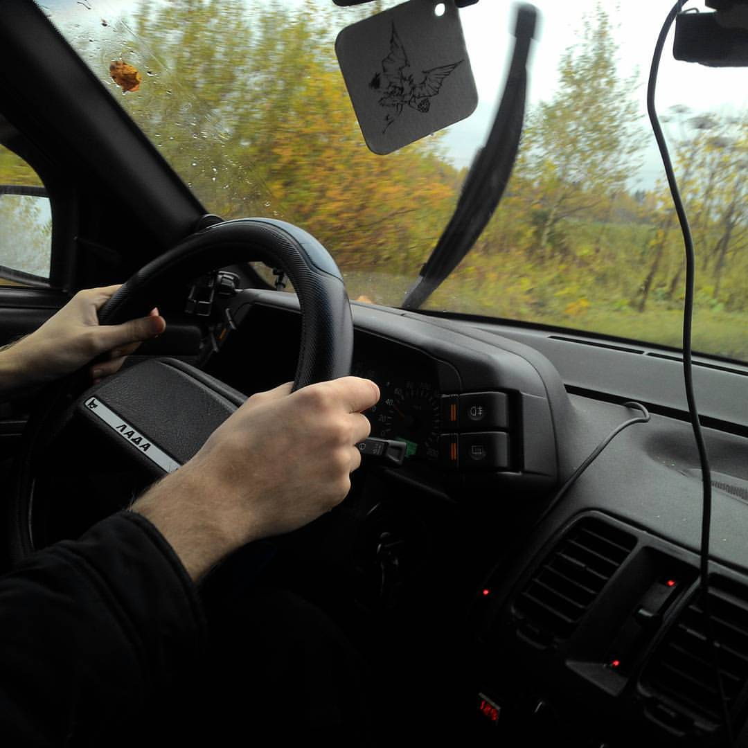 Вид из салона автомобиля ВАЗ (Lada): руки водителя в чёрной одежде держат рулевое колесо с надписью «ЛАДА»; через мокрое лобовое стекло с поднятыми дворниками видны осенние деревья с жёлто-зелёной листвой; с зеркала заднего вида свисает ароматизатор в форме тёмного мультперсонажа; интерьер тёмный