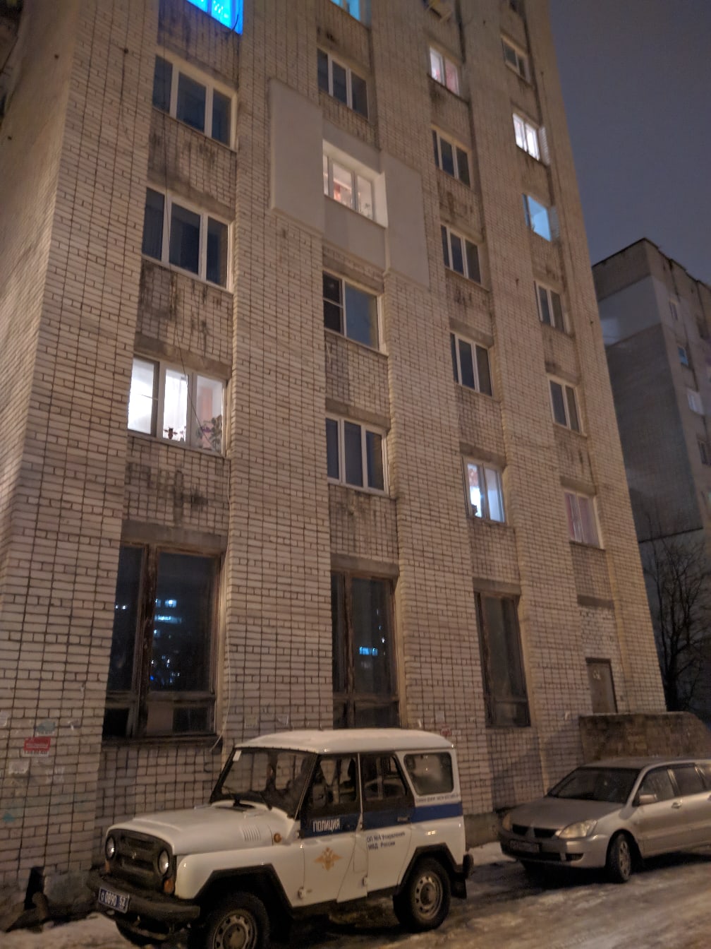 9-этажный панельный жилой дом ночью: светятся несколько окон, снег на земле; у подъезда припаркованы УАЗ-469 и легковой автомобиль