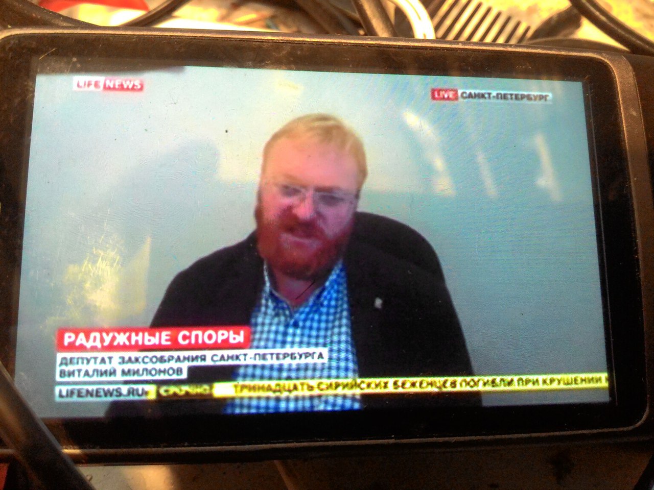 Экран смартфона-навигатора: LifeNews, LIVE Санкт-Петербург; заголовок «РАДУЖНЫЕ СПОРЫ»; подпись «Депутат Заксобрания Санкт-Петербурга Виталий Милонов»; бегущая строка «lifenews.ru ● СРОЧНО ТРИНАДЦАТЬ СИРИЙСКИХ БЕЖЕНЦЕВ ПОГИБЛИ ПРИ КРУШЕНИИ...»