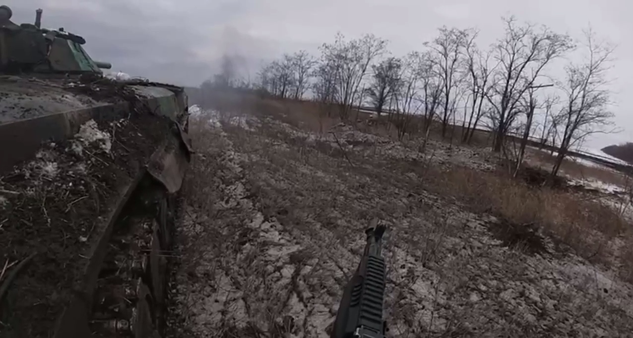 📷 Вид с борта военной бронемашины: камуфлированная бронетехника (БМП) в зимнем поле, на переднем плане ствол оружия, вдали голые деревья и дым.