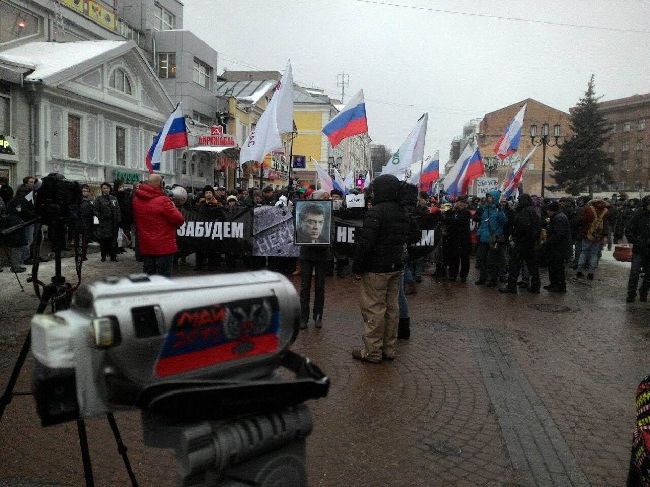 📷 Городская акция (митинг): группа людей с российскими триколорами и плакатами, среди которых виден плакат «НЕ ЗАБУДЕМ»; пасмурный зимний день; деревья без листьев; видеооператоры на переднем плане.