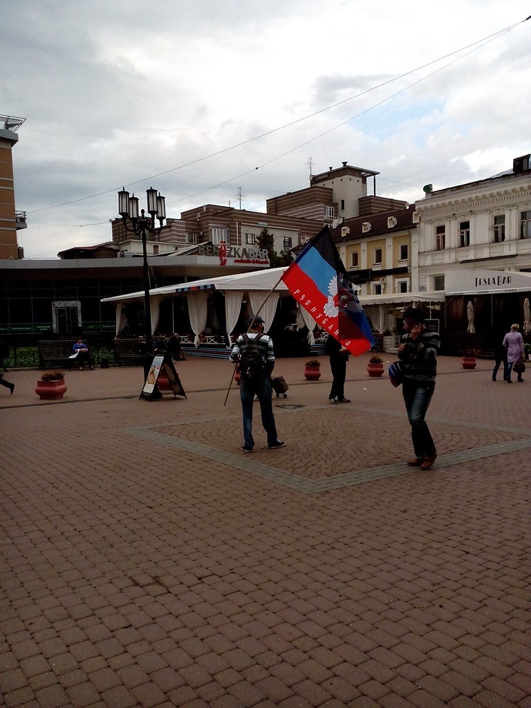 Городская площадь Нижнего Новгорода: двое мужчин держат флаг ДНР (чёрно-сине-красный с орлом); рестораны и исторические здания на заднем плане