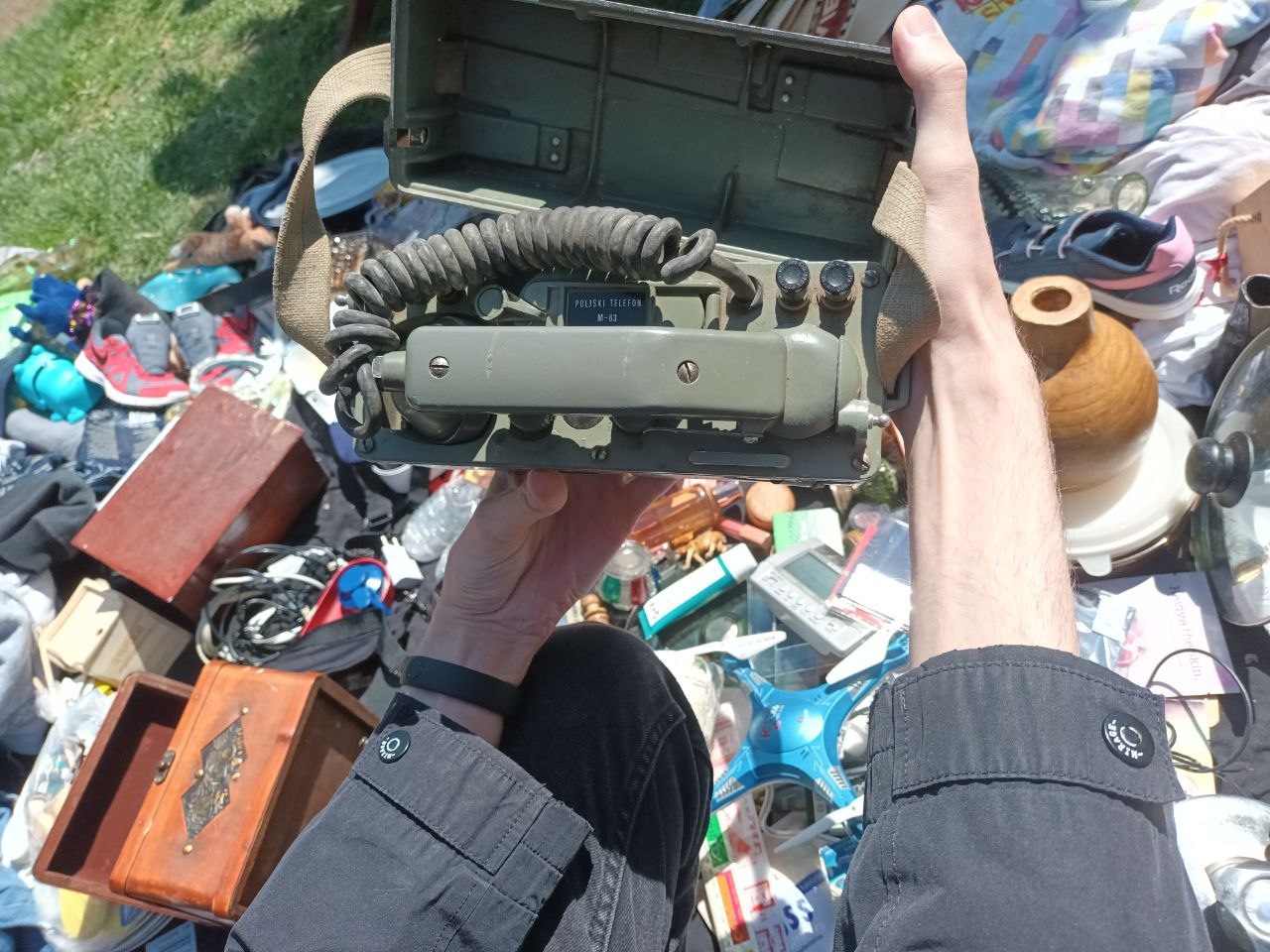 📷 Рука держит открытый военно-полевой телефон (зелёный корпус, витой шнур) над разложенными вещами на блошином рынке.