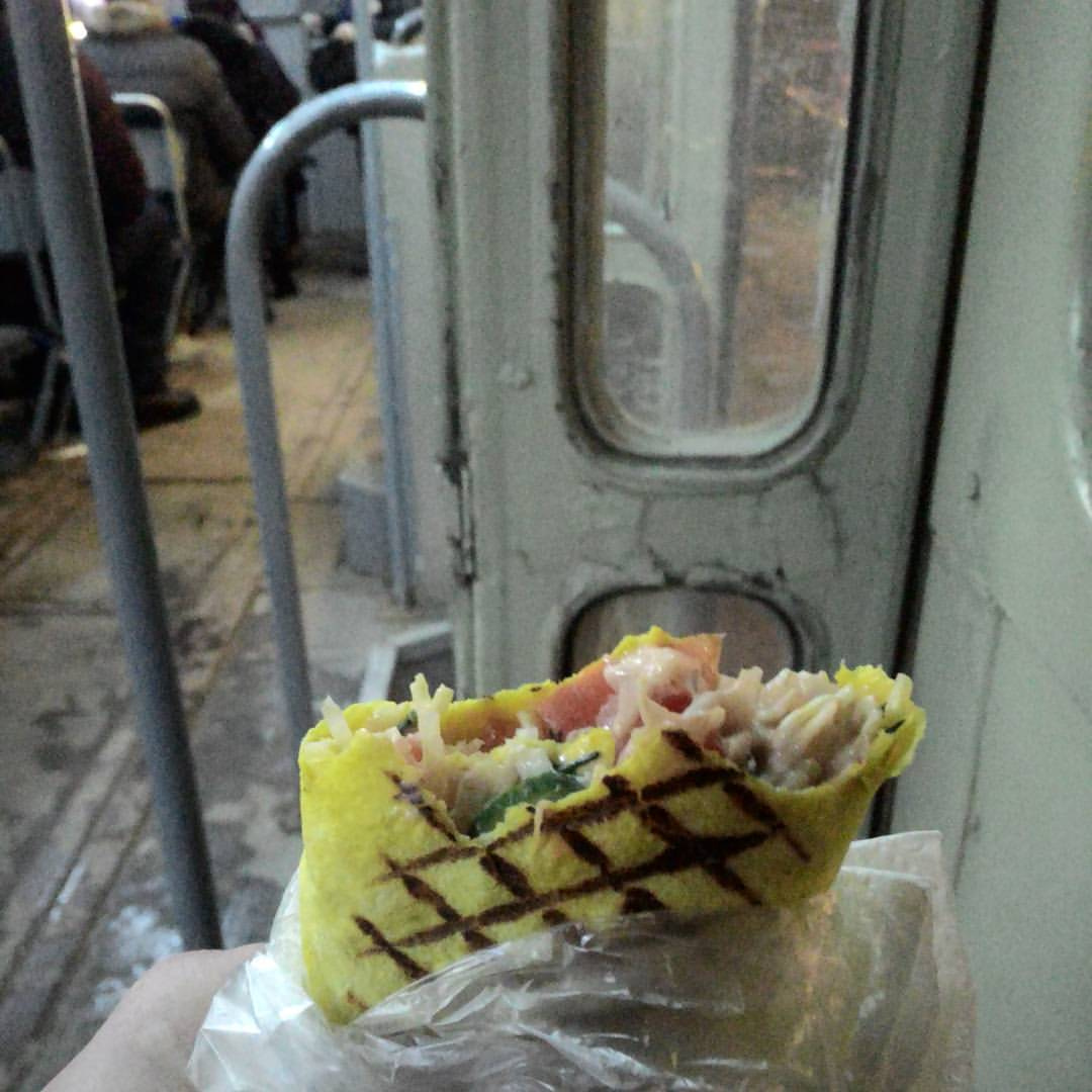 Крупный план шаурмы в руке: жёлтый лаваш с характерными гриль-полосками, начинка — курица, овощи (помидор, зелень), соус, в бумажной упаковке; на фоне — салон автобуса/троллейбуса (металлические поручни, дверь, сидящие пассажиры)