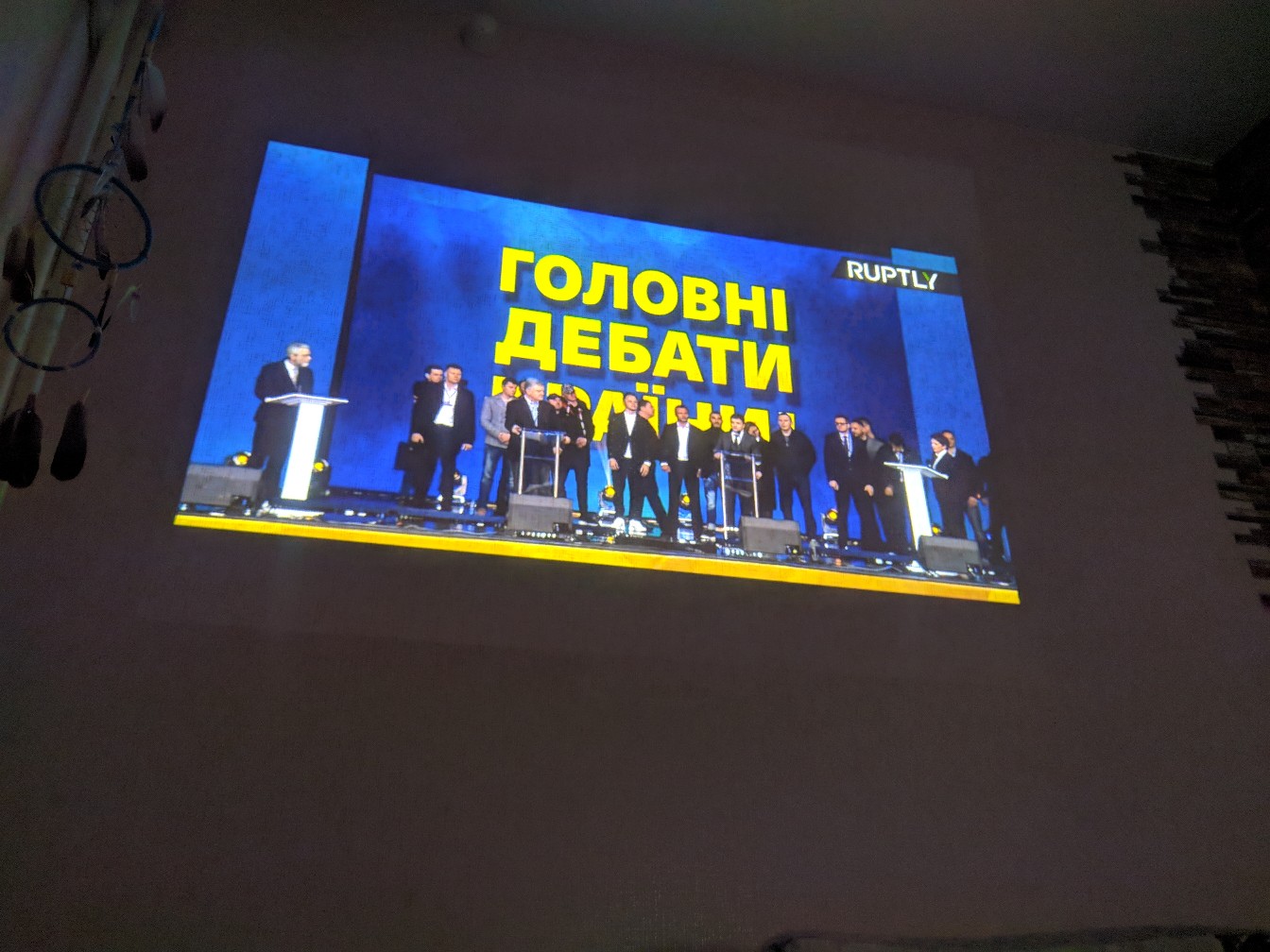 Проекция на стену тёмной комнаты — трансляция Ruptly: надпись «ГОЛОВНІ ДЕБАТИ [КРАЇНИ]» жёлтыми буквами на синем фоне; несколько участников дебатов на сцене со стойками для микрофонов