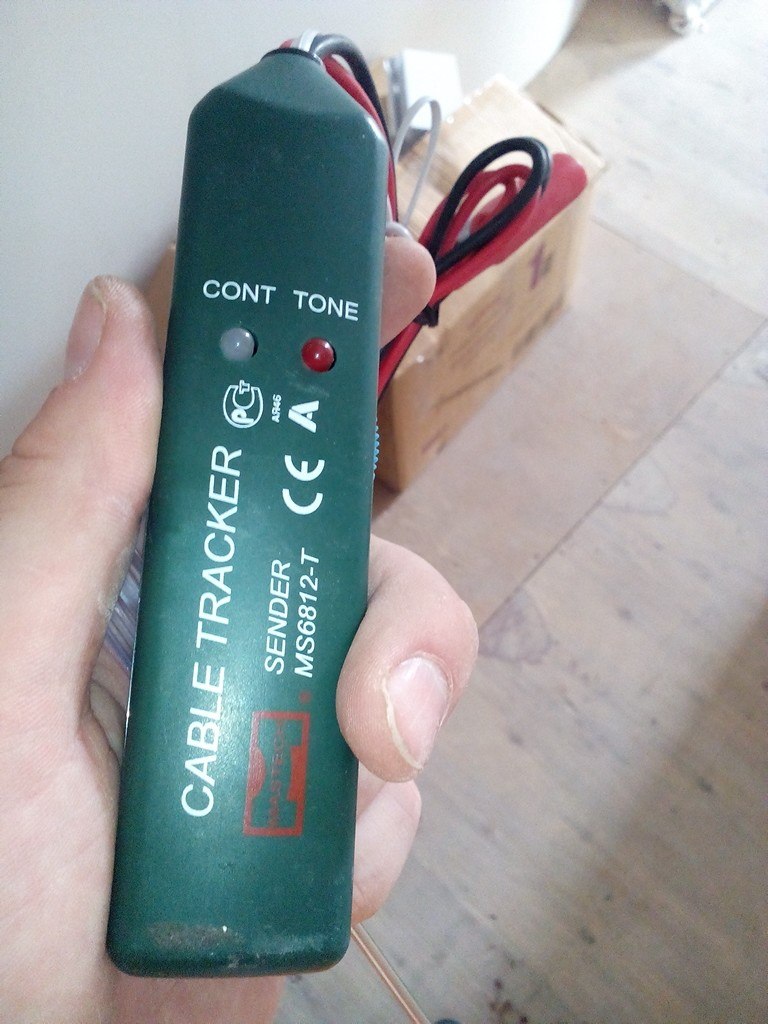 Зелёный Cable Tracker SENDER MS6812-T в руке, индикаторы CONT и TONE; на заднем плане коробка и провода