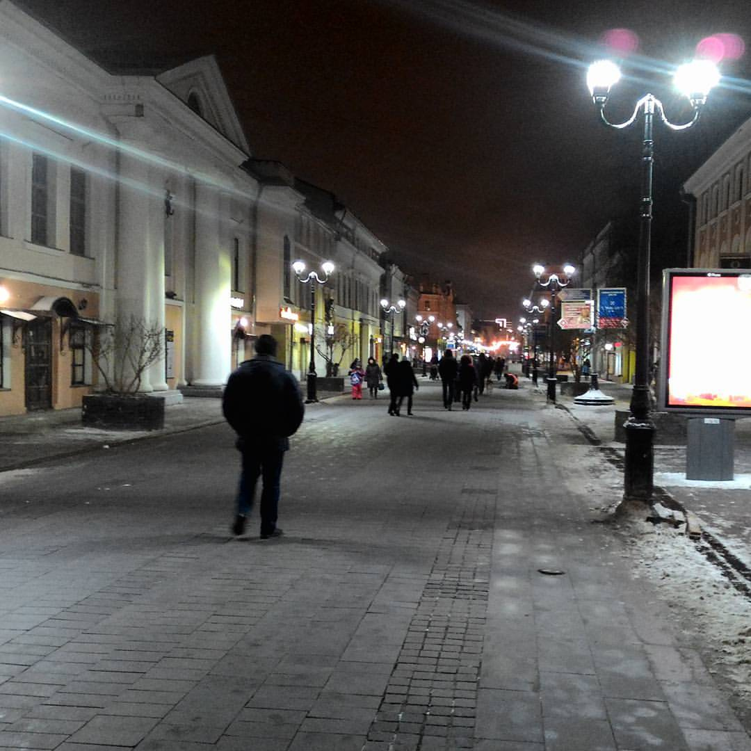 Ночная пешеходная улица в историческом центре города: широкий брусчатый тротуар, по бокам — классические здания с колоннами и фасадами; вдоль улицы — старинные фонарные столбы с круглыми плафонами; вдали — огни и прохожие; на земле — следы снега; справа — рекламный лайтбокс