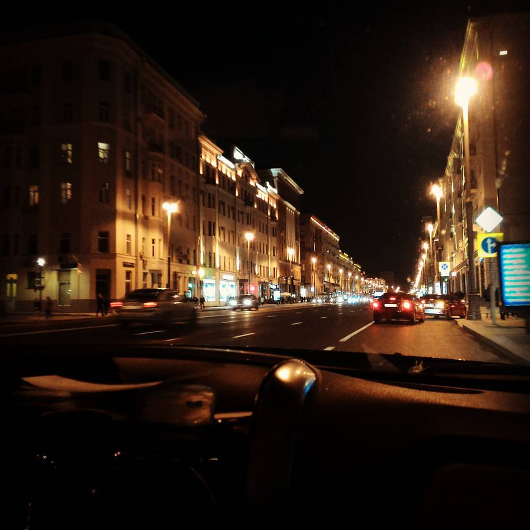 Вид через лобовое стекло автомобиля на широкий ночной московский бульвар: по обеим сторонам — многоэтажные здания сталинской/неоклассической архитектуры с тёплой подсветкой, несколько рядов движущихся автомобилей, уличные фонари; на правом краю — синий дорожный указатель; торпедо автомобиля видно снизу