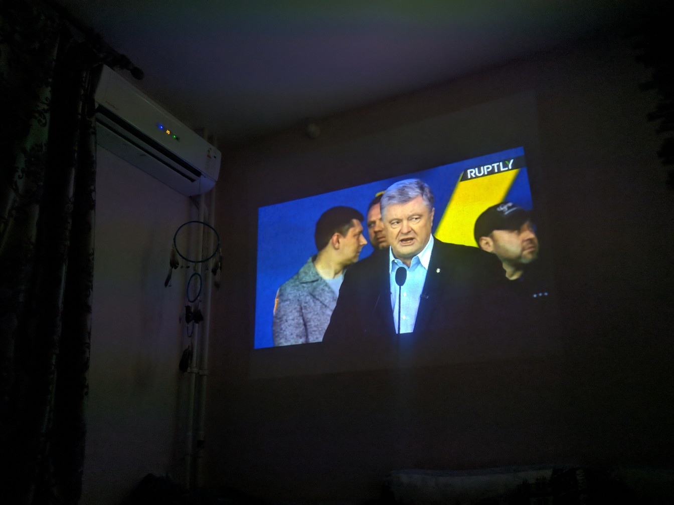 Проекция на стену тёмной комнаты — трансляция Ruptly: Порошенко в синей рубашке за микрофоном на сине-жёлтом фоне; кондиционер справа, ловец снов слева