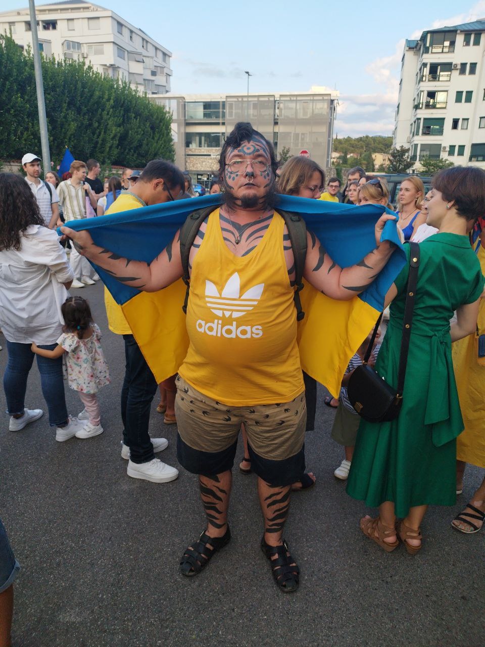 📷 Мужчина в жёлто-синей футболке adidas с украинским флагом за спиной, на лице — фурри-боевой грим (чёрно-бежевый окрас). На заднем плане — толпа людей на улице.