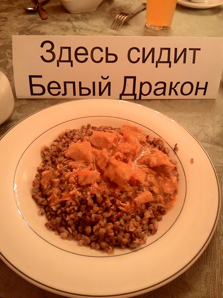 Белая тарелка с гречневой кашей и кусочками курицы в томатном соусе на столе в столовой; перед тарелкой стоит именная табличка «Здесь сидит Белый Дракон»; на заднем плане — стакан с соком и столовые приборы
