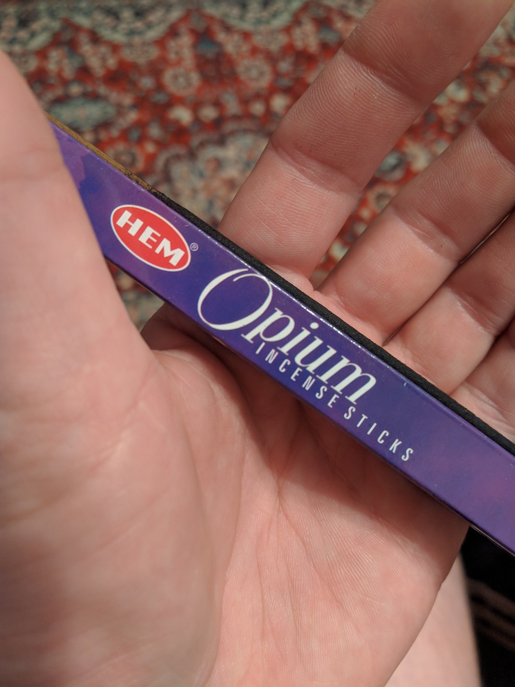 Рука держит фиолетовую упаковку «HEM® Opium Incense Sticks»; на фоне ковёр с узором
