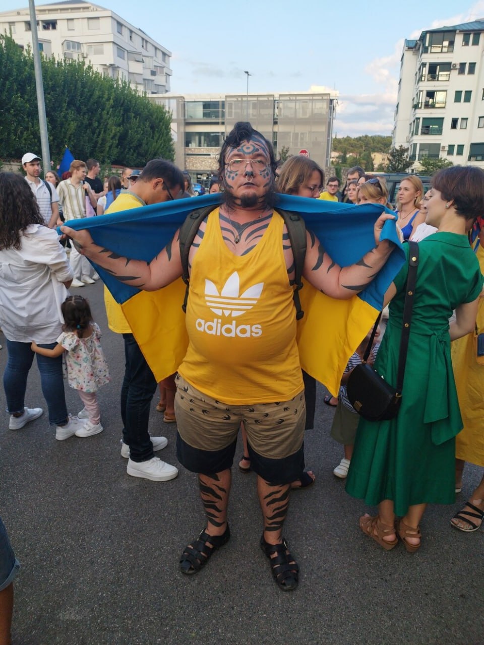 📷 Тот же мужчина в жёлто-синей майке Adidas и шортах, с флагом Украины за спиной; стоит в толпе на улице, городской фон — Черногория.