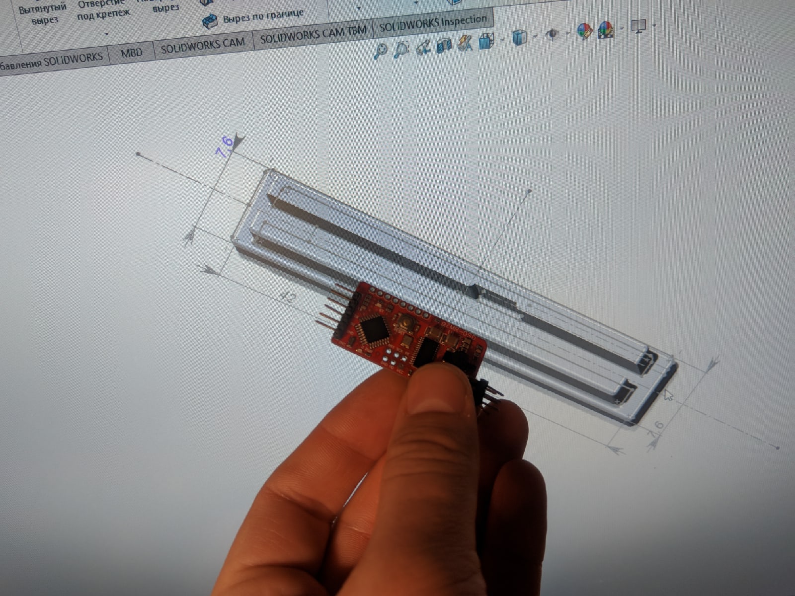 📷 Рука держит плату PCB с длинным чёрным компонентом и красно-оранжевыми проводами; на фоне — монитор с техническими схемами.