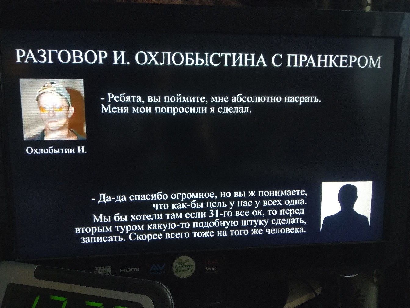 Экран телевизора с надписью «РАЗГОВОР И. ОХЛОБЫСТИНА С ПРАНКЕРОМ»: слева фото Охлобытина И. в очках-авиаторах и подпись «Охлобытин И.» — цитата «Ребята, вы поймите, мне абсолютно насрать. Меня мои попросили я сделал.»; справа чёрный силуэт пранкера — цитата «Да-да спасибо огромное, но вы ж понимаете, что как-бы цель у нас у всех одна. Мы бы хотели там если 31-го все ок, то перед вторым туром какую-то подобную штуку сделать, записать. Скорее всего тоже на того же человека.»