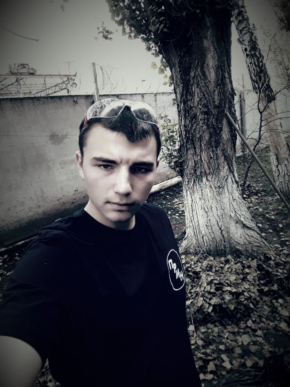 📷 Молодой мужчина в чёрной футболке, монохромное сэлфи на фоне дерева и кустарника.