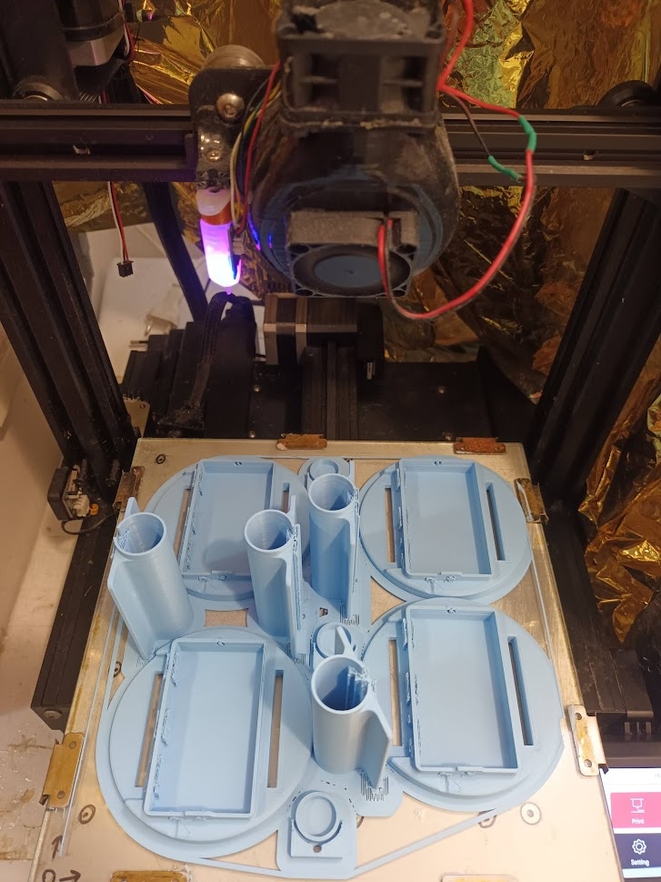 📷 3D-принтер печатает голубые пластиковые корпуса для SlimeVR-трекеров: прямоугольные коробочки с цилиндрическими стойками лежат на рабочей плите, сопло горит фиолетовым.