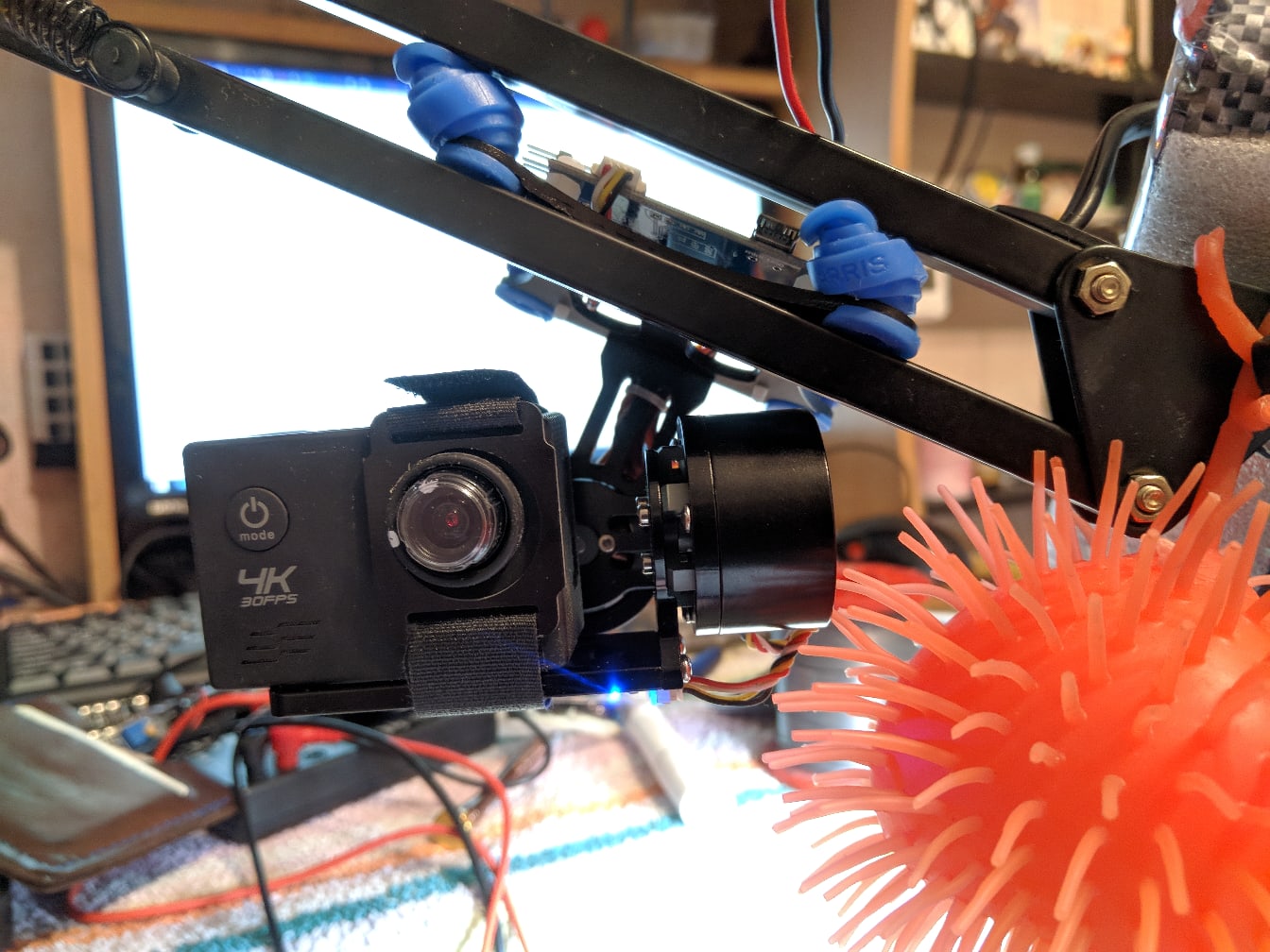 📷 Трёхосевой подвес (gimbal) с экшн-камерой с надписью «4K» на чёрной металлической раме дрона: синие провода, красный помпон справа, рабочий стол с монитором на заднем плане