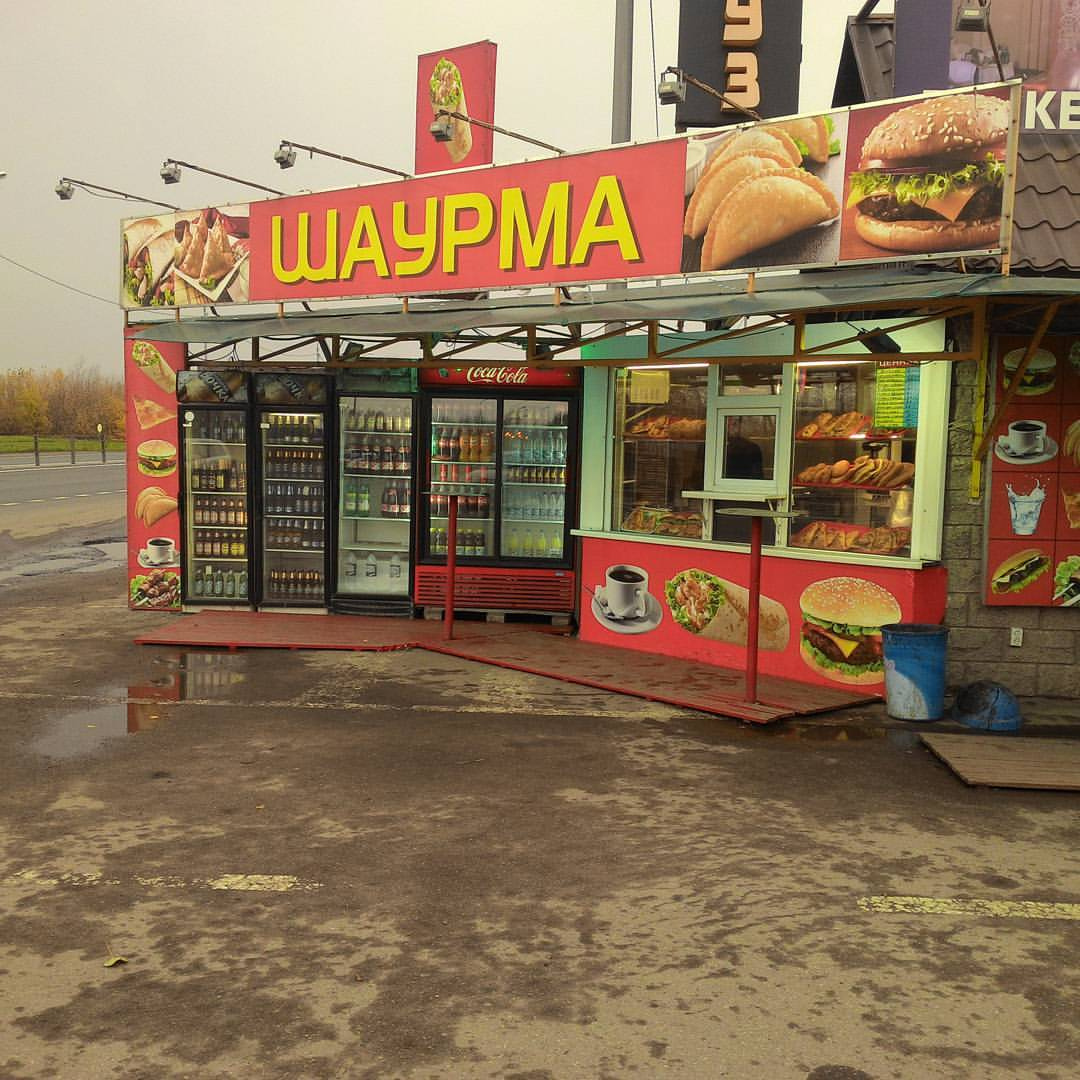 Придорожный ларёк с яркой жёлто-красной вывеской «ШАУРМА» на мокрой асфальтовой стоянке в пасмурный осенний день; в ларьке — витрины с едой, холодильники с напитками, фотографии блюд; стоянка пустая, мокрый асфальт
