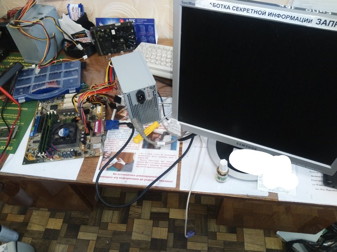 Компьютерный стол: материнская плата ASUS с процессором, блок питания, монитор Samsung — на заднем плане бумажка «РАБОТКА СЕКРЕТНОЙ ИНФОРМАЦИИ ЗАП[РЕЩЕНА]»