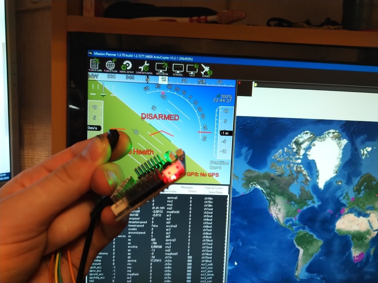 Рука держит плату ArduPilot с красными LED перед монитором с Mission Planner (надпись «DISARMED», искусственный горизонт); справа второй монитор с Google Maps