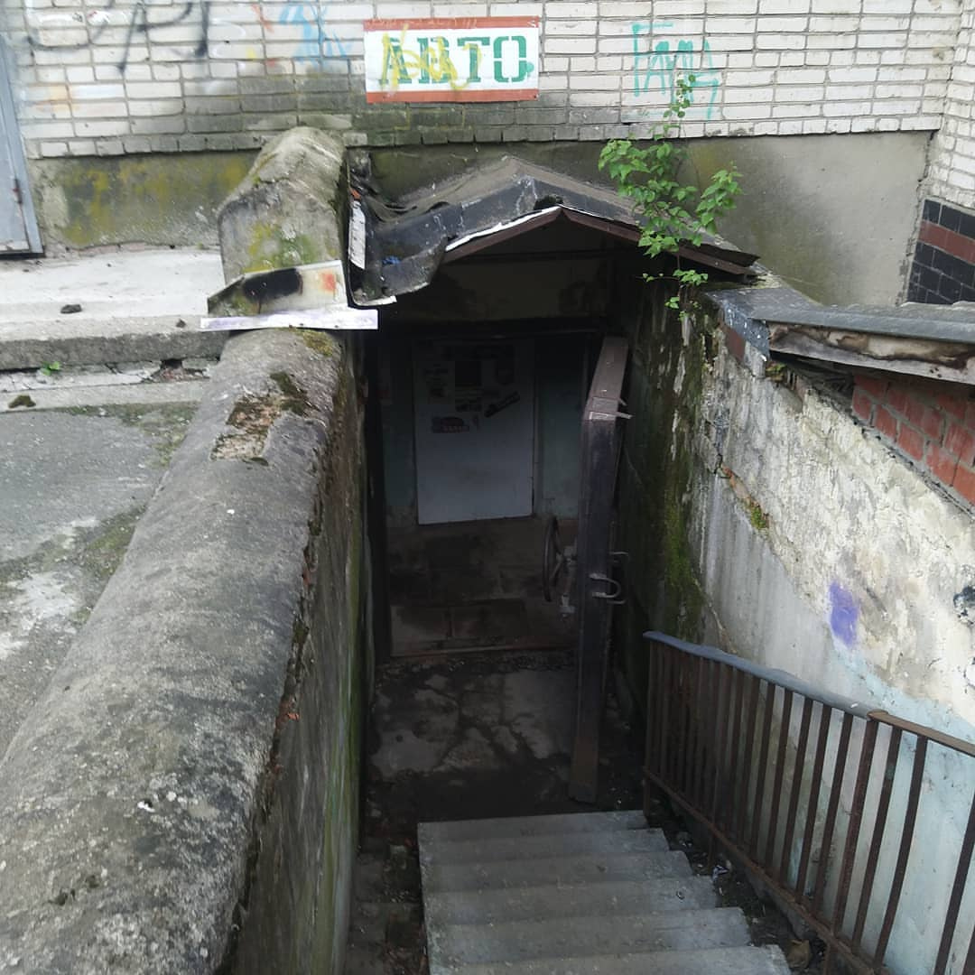 Вход в подвальный магазин автозапчастей: бетонные ступеньки ведут вниз к тёмному дверному проёму; над входом — вывеска «АВТО» зелёными буквами; облезлые кирпично-бетонные стены с граффити; металлические перила справа; над дверью — небольшой навес