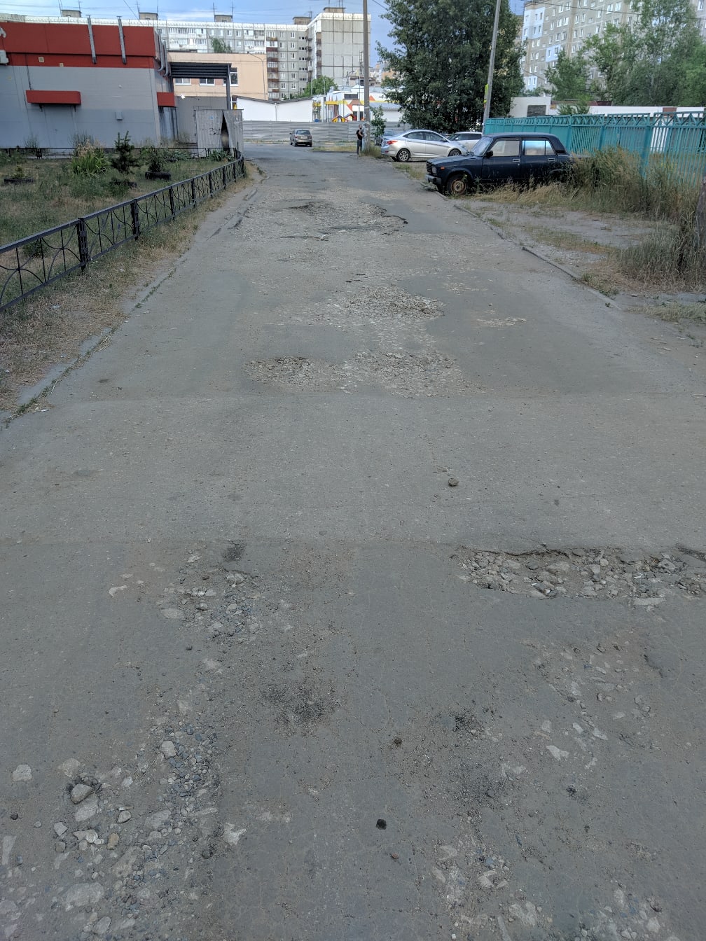 📷 Разбитый асфальт с ямами и трещинами на тротуаре, вдали — проезжая часть с припаркованными автомобилями и жилые дома