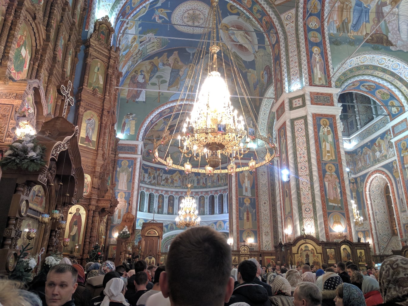 Интерьер православного храма полный прихожан: расписные своды и стены с фресками, большие хрустальные люстры, позолоченный иконостас, арочные проёмы с росписями